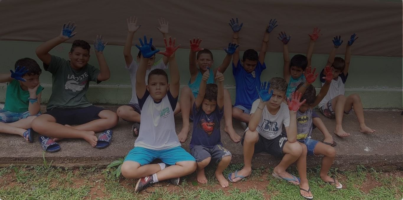 Crianças do Projeto Beija-Flor sentadas lado a lado, com as mãos pintadas de tinta levantadas